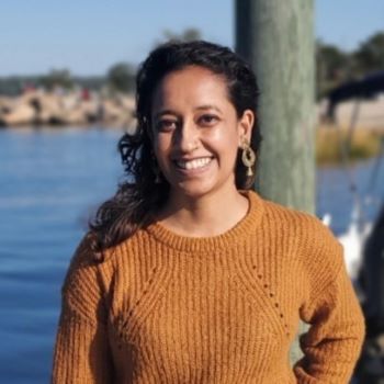 Harshita stands smiling in a orange sweater in front of a large body of water.