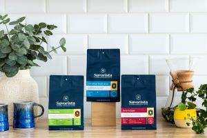 Three blue bags of coffee grounds sit on a light colored wood table in front of a white brick wall