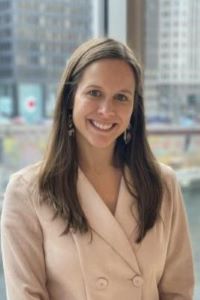 Photo of Ashely O'Brien OMBA student with long dark hair smiling and wearing a light colored suit 