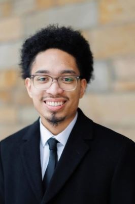 Photo of man with dark hair wearing a black suit with a white shirt and wearing glasses.