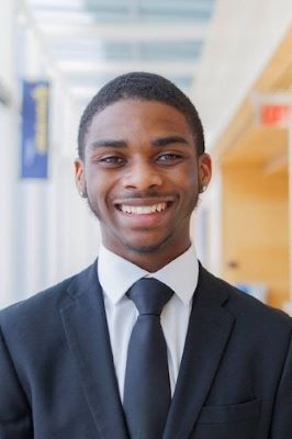 Photo of a man with short dark hair wearing a black suit with a white shirt and a black tie and smiling 