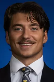 Man with dark hair and mustache wearing a dark suit with white shirt and Michigan tie