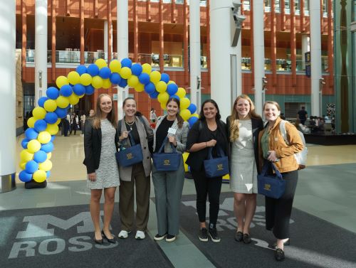 A group of six women smiling and holding tote bags standing in the Winter Garden with maize and blue balloons in the background