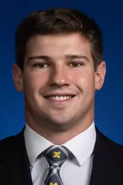 Man with dark hair wearing a dark suit with a white shirt and a Michigan tie