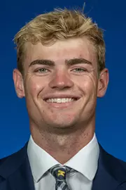 Man with blond hair and dark eyebrows wearing a dark suit with white shirt and Michigan tie