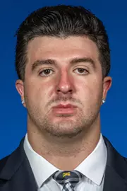 Man with dark hair wearing a dark suit and white shirt and Michigan tie