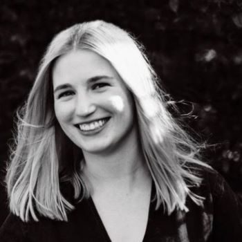 A black and white photo of Emily smiling, standing in front of the leaves of a tree or bush