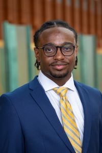 Photo of Evan Stocker Michigan Ross BBA student wearing a navy blue suit jacket with a yellow and blue striped tie. He has dark hair and is wearing glasses and smiling. 