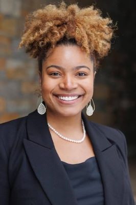 Photo of woman with light, curly hair smiling and wearing a black suit with a black shirt and long earrings