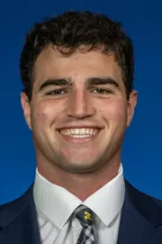 Man with dark hair wearing a dark suit with a white shirt with Michigan tie