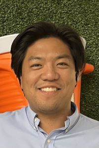 Photo of a man with dark hair smiling and wearing a light blue button up shirt