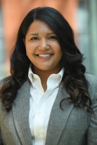 Photo of a woman with long dark hair smiling and wearing a gray suit with a white button up shirt