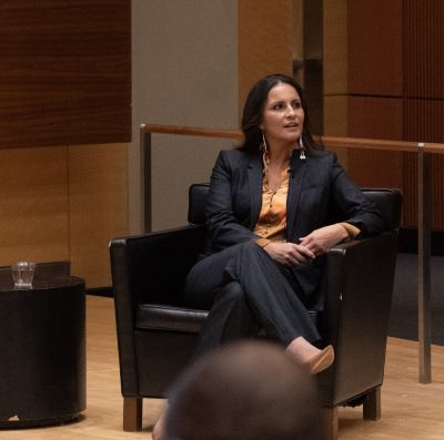 A woman in business attire sitting in a chair onstage speaking to the crowd.