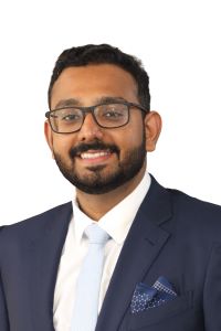 Photo of a man with dark hair smiling and wearing glasses with a navy blue suit with a white shirt and a light blue tie 