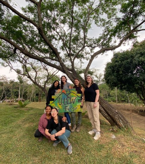 Students pose in the grass in front of a tree holding an ULEW Fuego sign