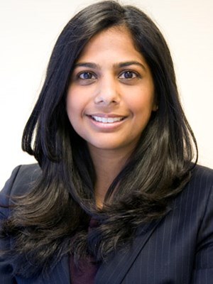 Saleena smiles, wearing a dark colored blazer with white stripes in front of a white background