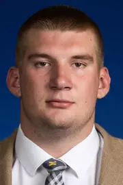 Man with short hair wearing a tan suit with a white shirt and Michigan tie