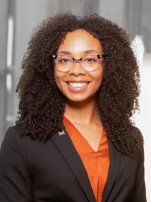 A woman with shoulder length brown hair smiling in a dark gray blazer with an orange shirt