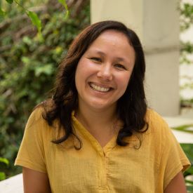 Carlotta smiles, wearing a yellow shirt and standing outside in front of a cement post and some plants