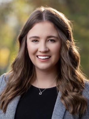 A woman with long brown hair wearing a gray blazer and black shirt smiles in a photo taken outside