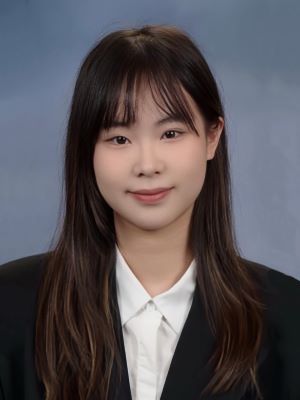 A woman with long brown hair wearing a black suit jacket and white shirt smiles in front of a greyish blue backdrop