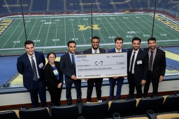 A group of seven graduate students standing in a box at the Big House overlooking the football field holding a giant check for $4000