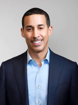 A man smiling in a dark blue jacket and blue dress shirt in front of a gray background