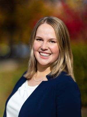 A woman with shoulder length blonde hair smiles, wearing a blue jacket and white shirt while standing outside near fall trees