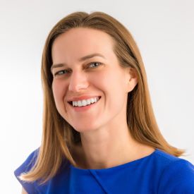 Kaitlin smiles, wearing a blue shirt and standing in front of a white background