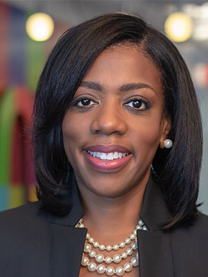 A woman with shoulder length dark brown hair smiling wearing a black blazer with a pearl necklace in front of a colorful background