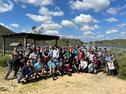 A large group of people pose on a sandy trail in front of a large body of water