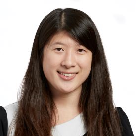 Natalie smiles, wearing a white and black shirt and standing in front of a white background