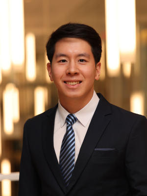 Prawonprat smiles in a black suit and blue tie in front of a blurred background of many bright lights