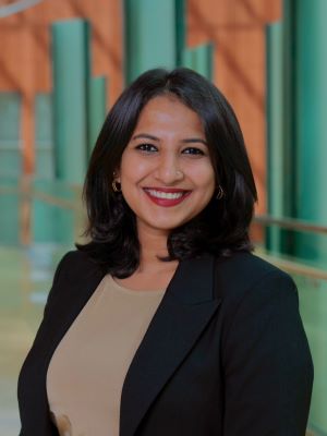 A woman with shoulder length dark brown hair smiles wearing a black blazer and brown shirt while standing in the Ross Winter Garden