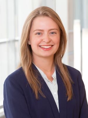 Rena smiles, wearing a blue blazer with a white shirt underneath. She is in the Ross building with the large windows behind her