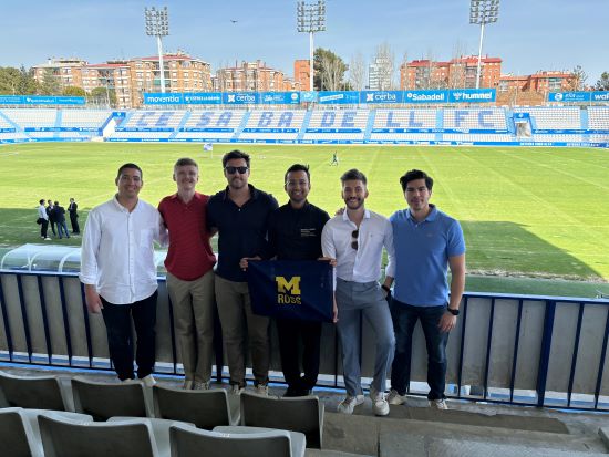 Students stand at the edge of the stands/seating area in front of a futbol field in the sun