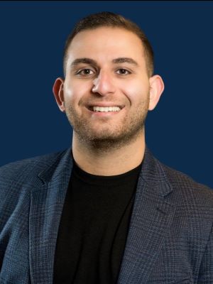 A man with short brown hair smiling, wearing a gray plaid blazer and black shirt in front of a dark blue background