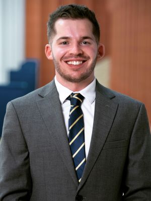 A man smiling with short brown hair and a dark gray suit with a white shirt and blue and gold striped tie