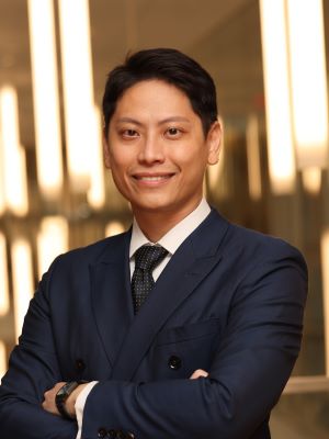 Steve Lin smiles for a photo in a navy blue suit and tie in front of a blurred background of many bright lights