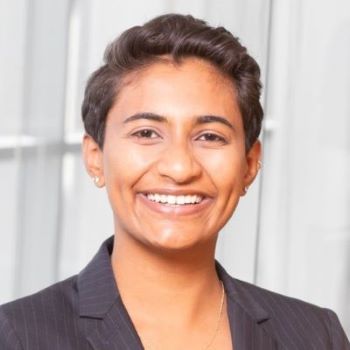 Shivali stands smiling in a gray blazer in the Ross Winter Garden in front of bright windows