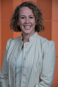 Woman with short curly brown hair wearing a white jacket and smiling 