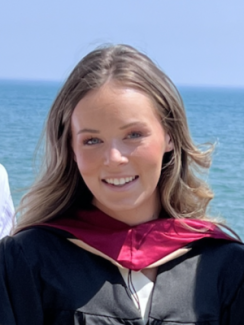 A woman with blonde hair smiles in a black jacket and pink hood, standing in front of the ocean or a lake.