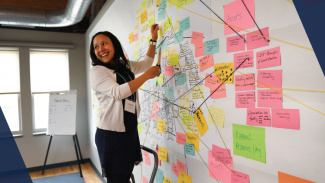 A woman places sticky notes on a large white wall, connecting the notes with tacks, string, and tape. She is in a well lit industrial room and is standing on a stepstool smiling.