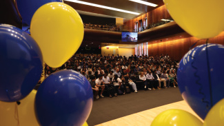 A crowd of students in the auditorium framed by maize and blue balloons in the foreground of the image