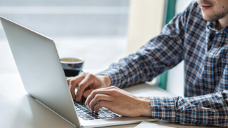 A man typing on a silver laptop computer in front of a window