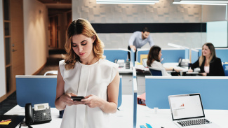 Young office worker staring at her phone, separated from colleagues gathering together without her in the background