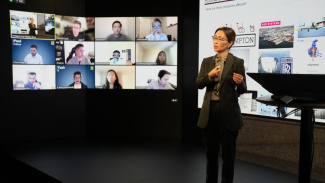 Photo of the Michigan Ross Digital Education Studio with a professor standing in the middle of the studio interacting with online students who are on a grid screen of 12 students. 