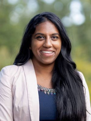 Supreya smiles, wearing a white/pale pink blazer over a blue shirt. Standing in front of many trees