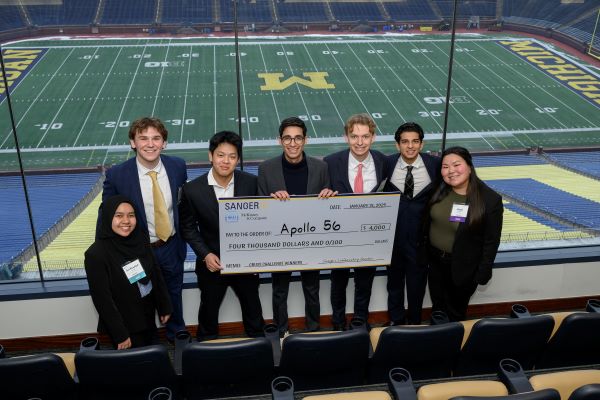 A group of seven undergraduate students standing in a box at the Big House overlooking the football field holding a giant check for $4000