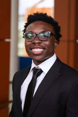 Photo of man with dark hair smiling and wearing glasses with a black suit and white shirt with a black tie